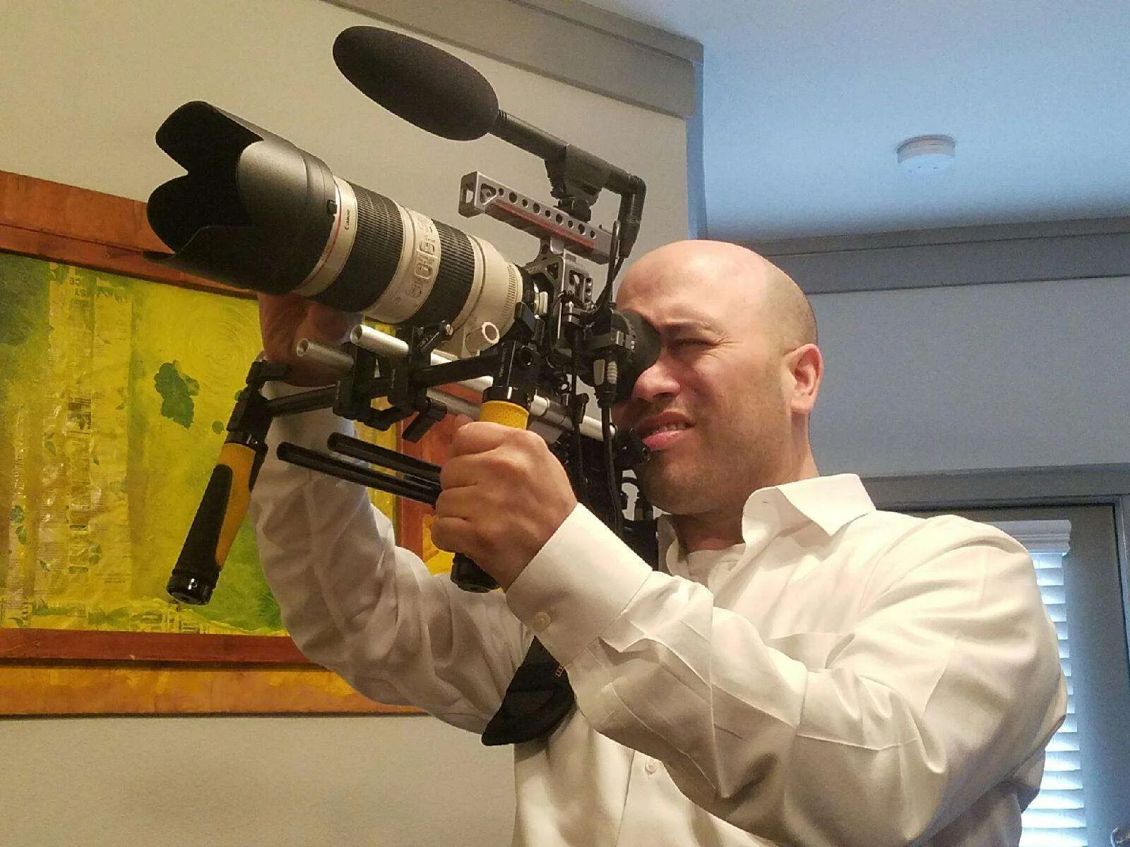 cameraman aiming a professional video camera with a large lens and attached microphone indoors, wearing a white shirt.