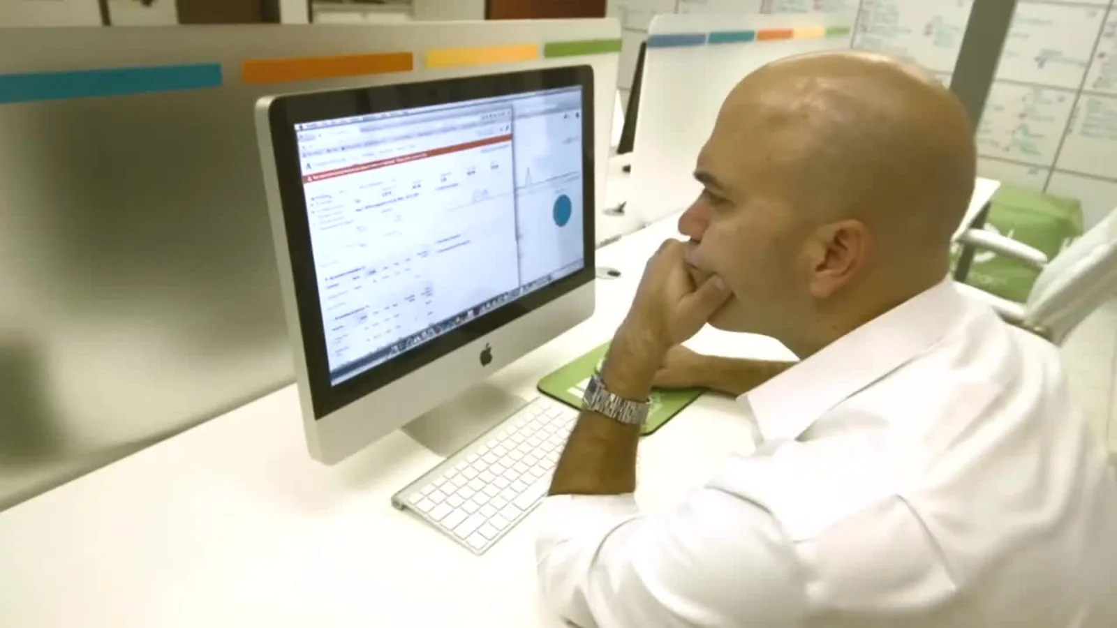 man in a white shirt sits at a desk and studies a large imac screen, chin resting on his hand in an office setting.