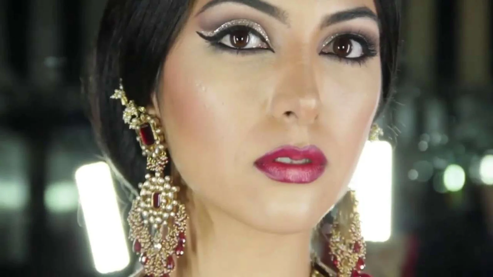 close-up of a woman wearing dramatic makeup with winged eyeliner and red lipstick, ornate gold earrings with red stones.